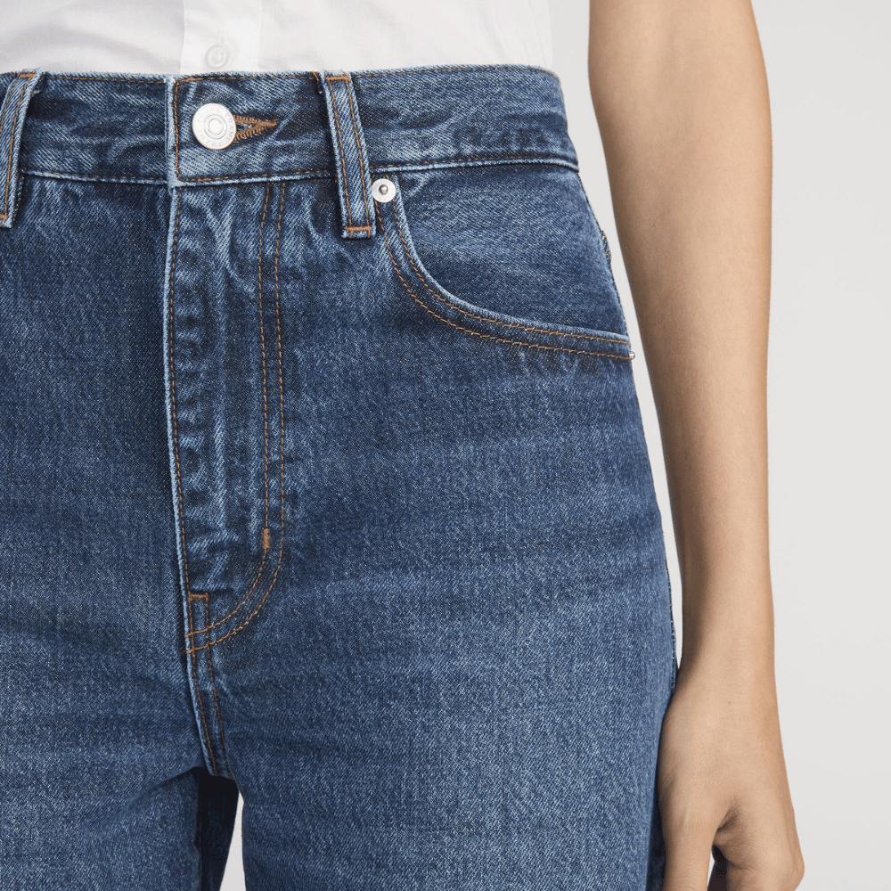 Close-up of a person wearing FRAME The Bow Jeans in Acumen New Fray at Riada Concept. High-rise rigid denim with a sculpted barrel leg and raw frayed hem -  with visible waistband, belt loops, front pocket, and stitching