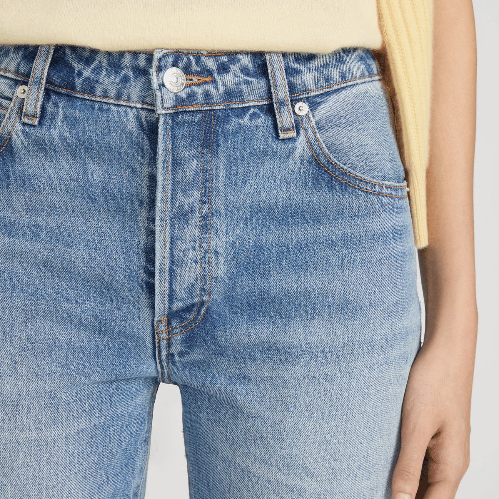 Close-up of FRAME The Essential Jean in Rapture at Riada Concept. Mid-rise full-length straight jeans in rigid regenerative cotton denim - paired with a pale yellow top. Focus on the waistband, front pockets, and buttons