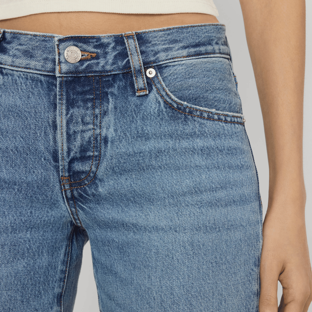 Close-up of FRAME The Gray Jean in Rapture at Riada Concept. Low-rise rigid denim with a long wide-leg flare, button fly and 34-inch inseam - showing the front waistband, button, belt loop, and pocket.